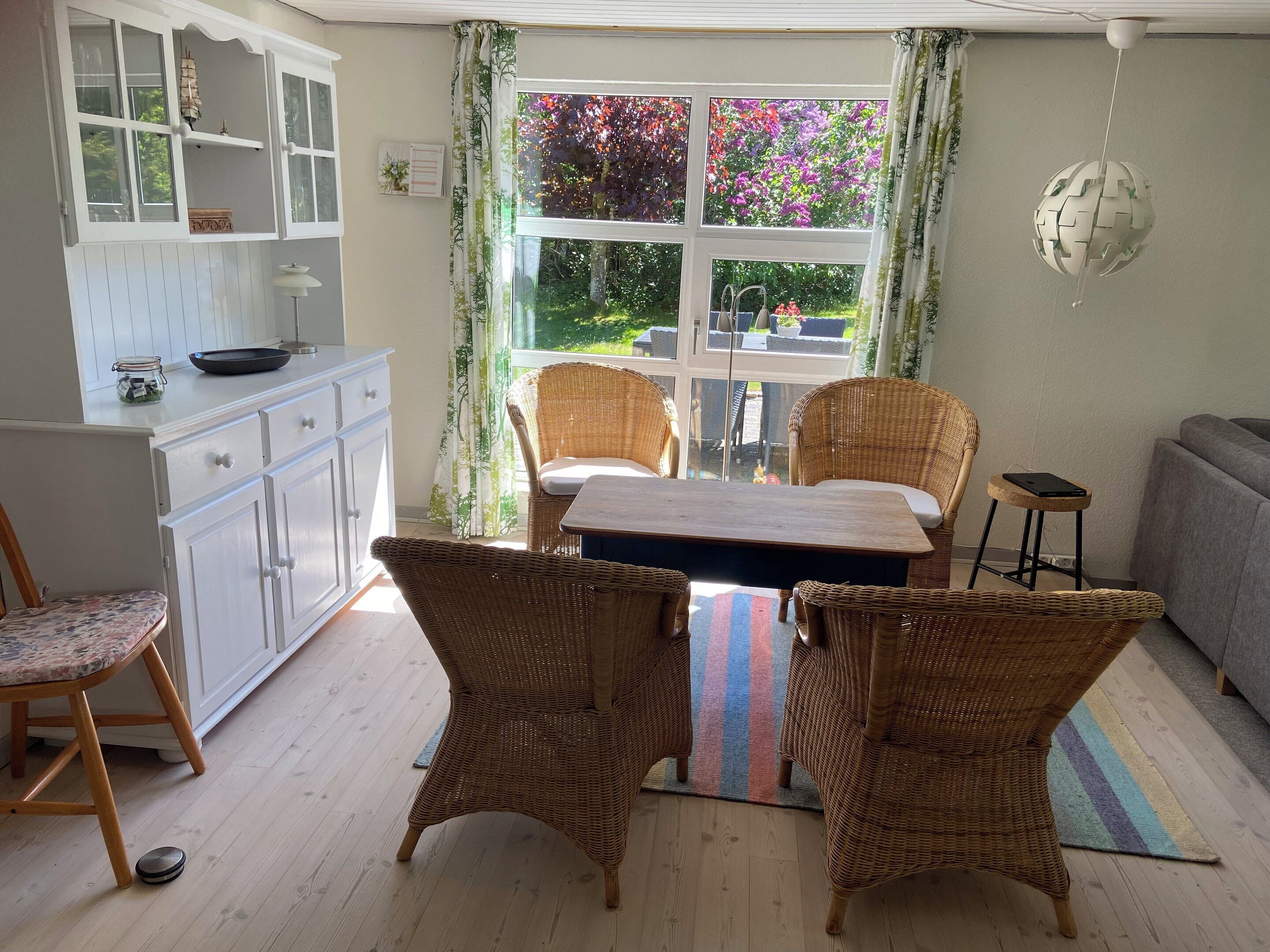 A room with a dining table, wicker chairs, a grey sofa, a small stool, a white cabinet, and a window with a view of greenery.