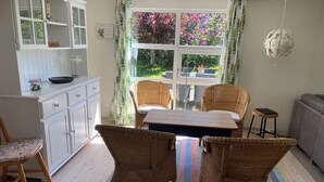 A room with a dining table, wicker chairs, a grey sofa, a small stool, a white cabinet, and a window with a view of greenery. - Holiday house – located in a forest area (Glesborg)