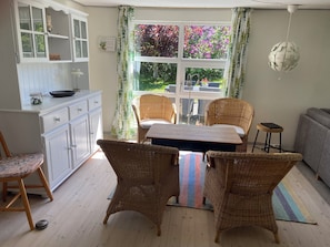 A room with a dining table, wicker chairs, a grey sofa, a small stool, a white cabinet, and a window with a view of greenery. - Holiday house located in a forest area (Glesborg)