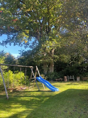 A playground with a slide and swings, surrounded by trees and grass. - Holiday house located in a forest area (Glesborg)