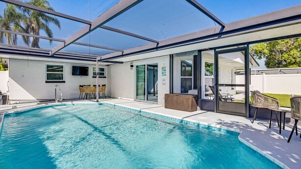 Ein Schwimmbecken mit Glasüberdachung, Sitzgelegenheiten im Freien und ein weißes Gebäude mit großen Fenstern.