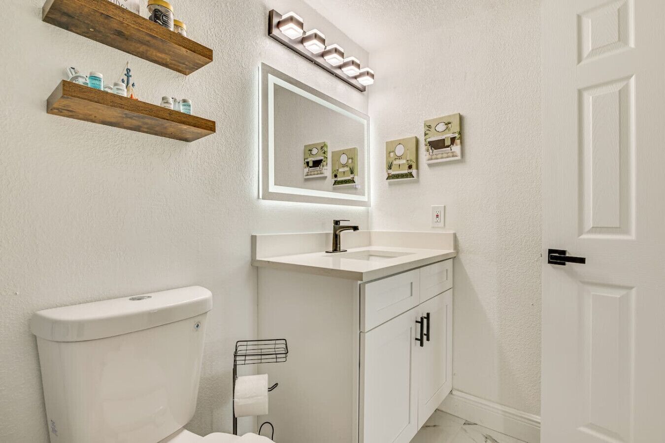 Une salle de bain avec des toilettes blanches, un lavabo avec un miroir au-dessus, et des étagères en bois au-dessus des toilettes.