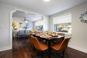 Uma zona de refeições com chão de madeira escura, uma mesa posta para quatro pessoas e vista para o exterior através de uma janela com persianas.