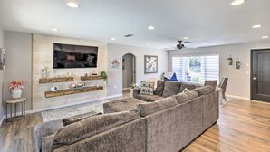 Uma sala de estar espaçosa com um amplo sofá modular, uma TV de tela plana fixada em uma parede de madeira e um ventilador de teto.