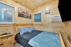 A wooden cabin room with a bed, a nightstand, and two windows with blinds.