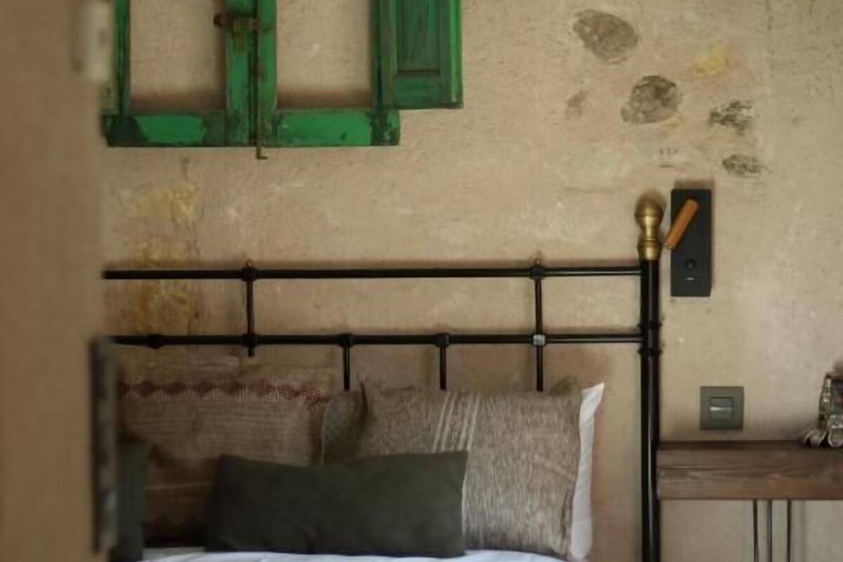 Een slaapkamer met een bed, kussens, een groen luik en een geweven lamp.