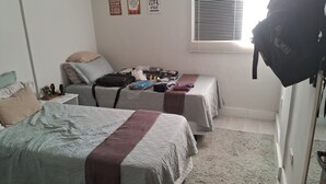 A bedroom with two beds, a window with blinds, and a suitcase on one bed. - Apartment for 4 people - Spacious and 700m from the beach. (João Pessoa)