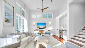A spacious living room with a high ceiling, a ceiling fan, and a large window with a view of the beach. There is a kitchen area with wooden cabinets and a staircase leading to another level. - Sol y Sombra (Santa Rosa Beach)
