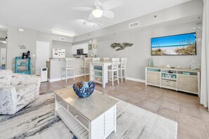 Un salon moderne avec un coin repas, une cuisine en arrière-plan, un ventilateur de plafond et une décoration murale sur le thème de la plage.