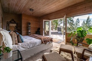 Quarto de cabana aconchegante com cama, uma pequena mesa e uma cadeira. Há uma estante, um espelho e uma janela com vista para árvores.