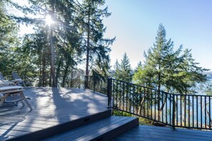 Um deck de madeira com mobiliário para áreas externas e guarda-corpo, com vista para um corpo d'água e uma floresta.