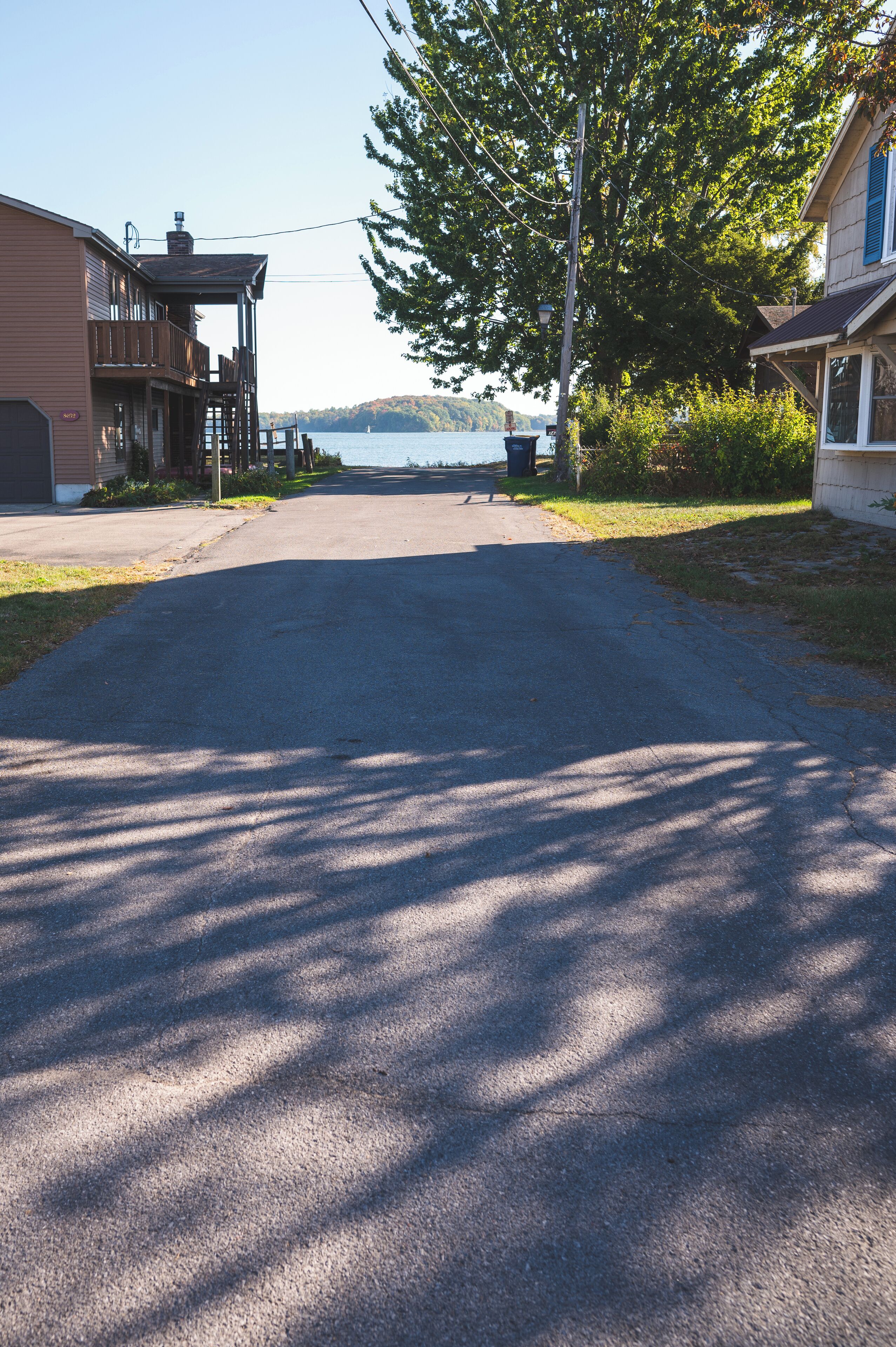 A residential street with houses on either side, a clear sky, and a body of water in the distance.