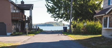 A residential street with houses on either side, a clear sky, and a body of water in the distance.