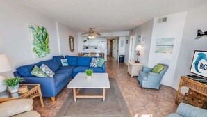 A living room with a blue sectional sofa, a white coffee table, and a blue armchair. There is a kitchen in the background with a ceiling fan. - New Private 4BR Ashworth,  Direct Oceanfront Views (North Myrtle Beach)