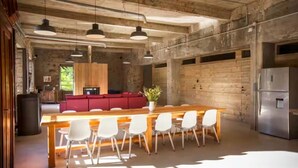 Un grand espace à manger avec une longue table en bois et des chaises blanches, un canapé rouge en arrière-plan, et un coin cuisine équipé d’appareils électroménagers en acier inoxydable.