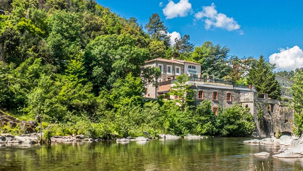 Un bâtiment en pierre doté de nombreuses fenêtres et d’un balcon, entouré d’une végétation luxuriante et bordé d’un lac aux eaux limpides.