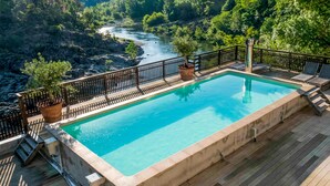 Une piscine rectangulaire dotée d’une terrasse en bois et d’une rambarde en métal, entourée d’une végétation luxuriante et bordée par une rivière en arrière-plan.