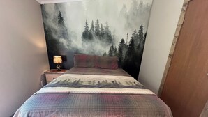 A bedroom with a bed, a nightstand with a lamp, and a forest-themed wall mural.