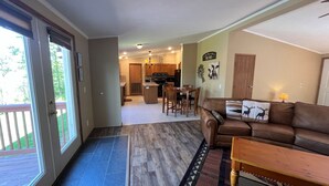 A spacious living area with a brown leather couch, a wooden coffee table, and a dining table with chairs. A kitchen is visible in the background with wooden cabinets and a built-in oven.