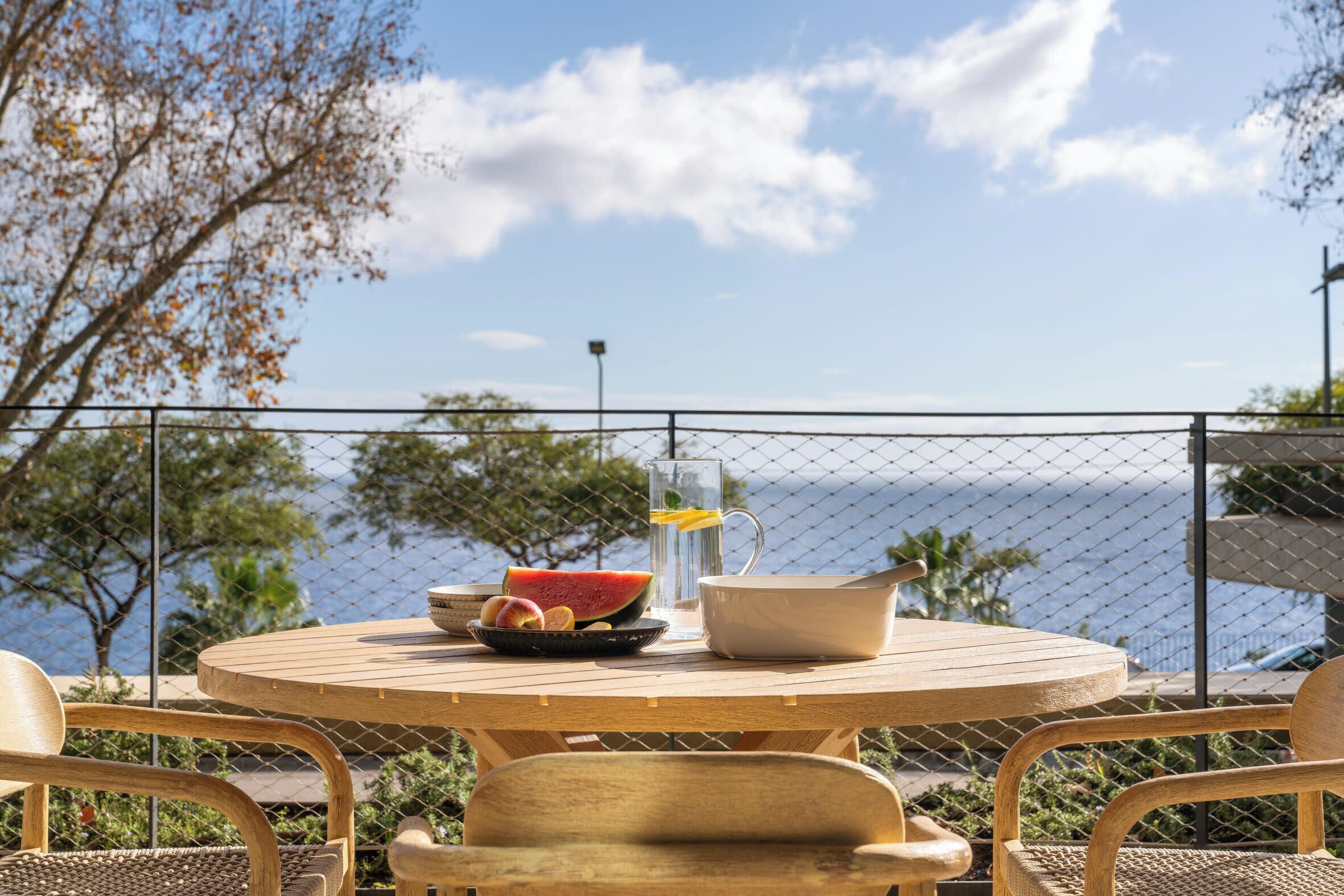 A round wooden table with a bowl of fruit, a pitcher, and a cup, set against a scenic ocean view with a fence in the foreground.