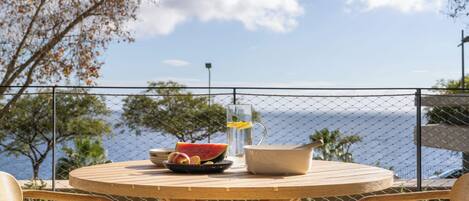 A round wooden table with a bowl of fruit, a pitcher, and a cup, set against a scenic ocean view with a fence in the foreground.