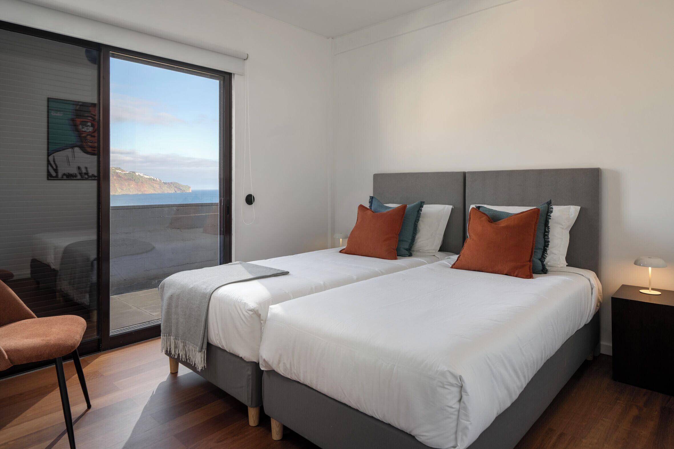 A modern bedroom with two beds, a sliding glass door leading to a balcony, and a small bedside table with a lamp.
