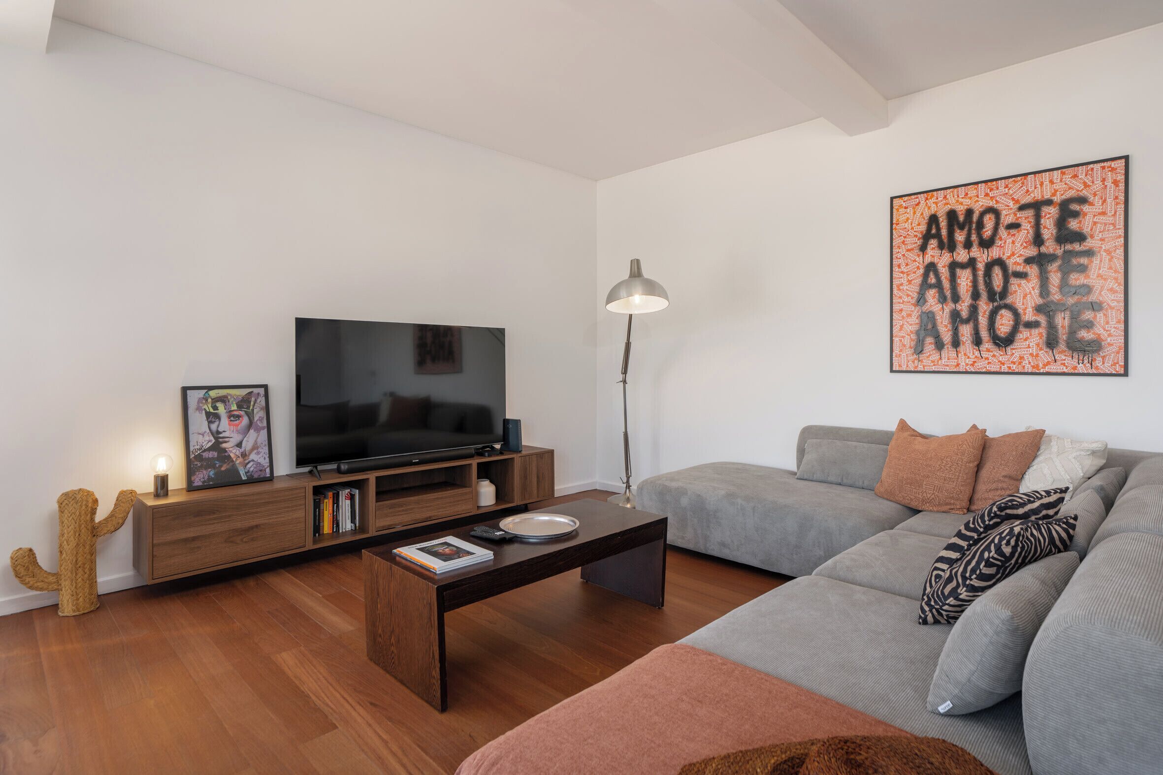 A modern living room with a flat-screen TV, wooden coffee table, grey sofa, and a cactus-shaped lamp.