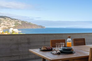 A table set with plates, glasses, a bottle of orange juice, and pastries, overlooking a coastal view. - Monumental Alta Vista – Panoramic View Balcony (São Martinho)