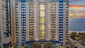 Um hotel com vários andares, átrio central, varandas em cada andar e vista para o oceano ao pôr do sol.