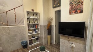 Een moderne woonkamer met een houten vloer, een glazen salontafel met documenten, een boekenkast, een flatscreen-tv aan de muur en een decoratief kunstwerk van touw.
