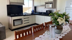 A modern kitchen with light wood cabinets, a black countertop, a built-in microwave, and a dining area with a wooden table and chairs. - Cozy beachfront apartment in Enseada, ideal for families and groups. (São Francisco do Sul)
