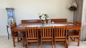 A dining table with six wooden chairs, a water dispenser, a vase with flowers, and a lace tablecloth. - Cozy beachfront apartment in Enseada, ideal for families and groups. (São Francisco do Sul)
