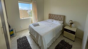 A bedroom with a bed, a nightstand, a lamp, a window with curtains, a ceiling fan, and a door. - Cozy beachfront apartment in Enseada, ideal for families and groups. (São Francisco do Sul)