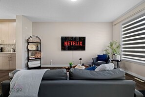 A modern living room with a grey sofa, a coffee table, and a TV mounted on the wall displaying 'NETFLIX'. - Large Home | Sleeps 12 | Yard | Near DT & Stadium (Edmonton)