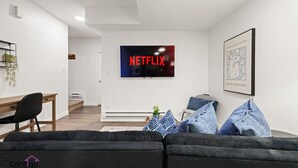Moderni olohuone, jossa on musta sohva, puinen työpöytä ja seinälle asennettu taulutelevisio, jonka näytöllä näkyy teksti ”NETFLIX”.