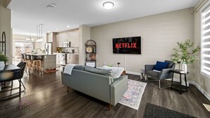 Moderni olohuone, jossa on harmaa sohva, sohvapöytä ja seinälle asennettu televisio, jonka näytöllä näkyy teksti ”NETFLIX”.