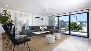 Una sala de estar moderna con sofá, sillón, mesa de centro y otomanas. Hay un ventanal con vistas a edificios y árboles.