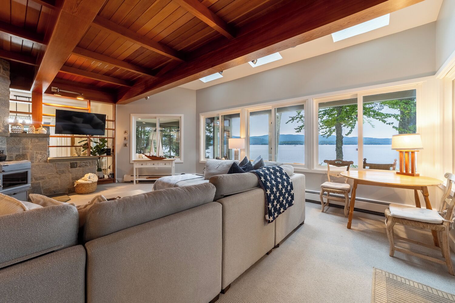 A modern living room with a large sectional sofa, a dining table with chairs, a fireplace, and large windows overlooking a body of water.