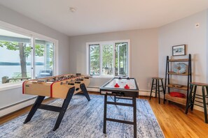 A room with two foosball tables, a wooden floor, a blue rug, and a window with a view of trees and a lake.