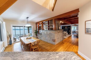 A spacious dining area with a large wooden table and chairs, a stone island with shelves, and a view of the sea through large windows.