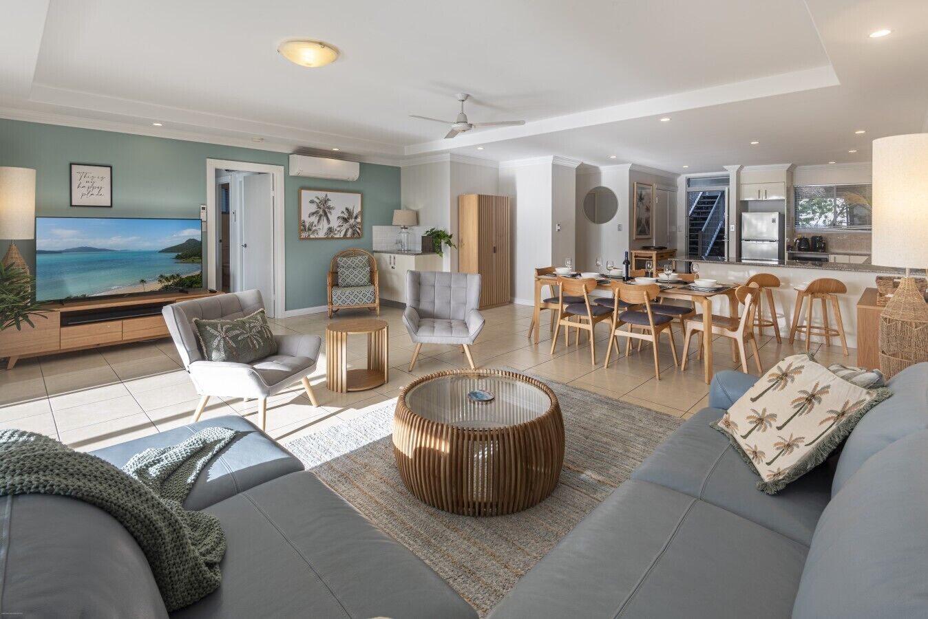 A modern living room with a large TV, comfortable seating, a dining area, and a kitchen.