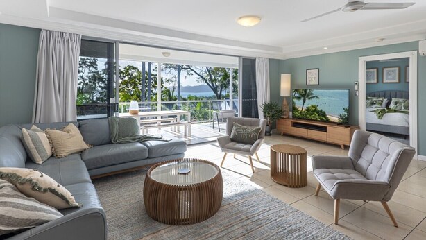 Ein modernes Wohnzimmer mit grauem Sofa, rundem Couchtisch und Sesseln. Ein Fernseher, eine Pflanze und ein Blick ins Freie sind zu sehen.