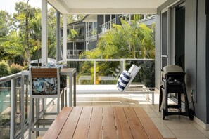A balcony with a wooden table, a cushioned chair, a grill, and a view of a multi-story building and surrounding greenery. - Blue Water Views 3 & Free Buggy- Hamilton Island (Hamilton Island)