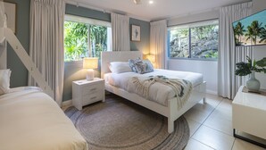 Ein Schlafzimmer mit Bett, Nachttisch und Beistelltisch, großen Fenstern mit Vorhängen, einem Deckenventilator und Blick ins Grüne.