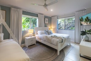 A bedroom with a bed, nightstand, and bedside table, large windows with curtains, a ceiling fan, and a view of greenery outside. - Blue Water Views 3 & Free Buggy- Hamilton Island (Hamilton Island)