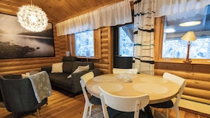 Ein gemütliches Blockhaus-Interieur mit einem runden Holztisch, weißen Stühlen, einem grauen Sofa und einer hängenden Leuchte.