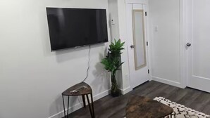 A living room with a flat-screen TV mounted on the wall, a small wooden stool, a potted plant, and a patterned rug on the floor. - Oshmild Suites, 5 mins to various shops and Cinema (Saskatoon)