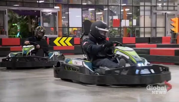 Two people in go-karts inside a building with glass windows and red seating.