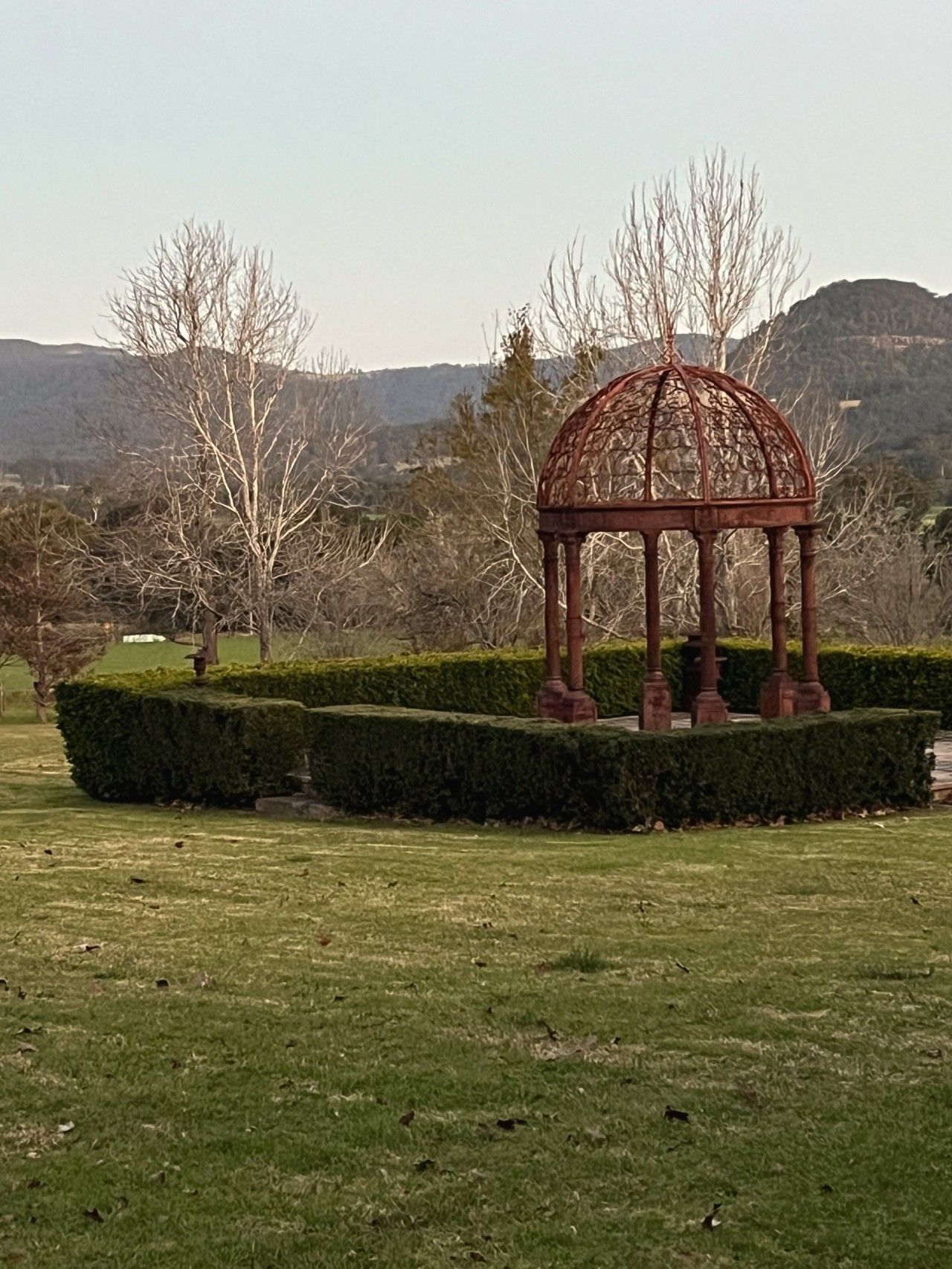Una glorieta en un parque con un seto cuidado y montañas al fondo.