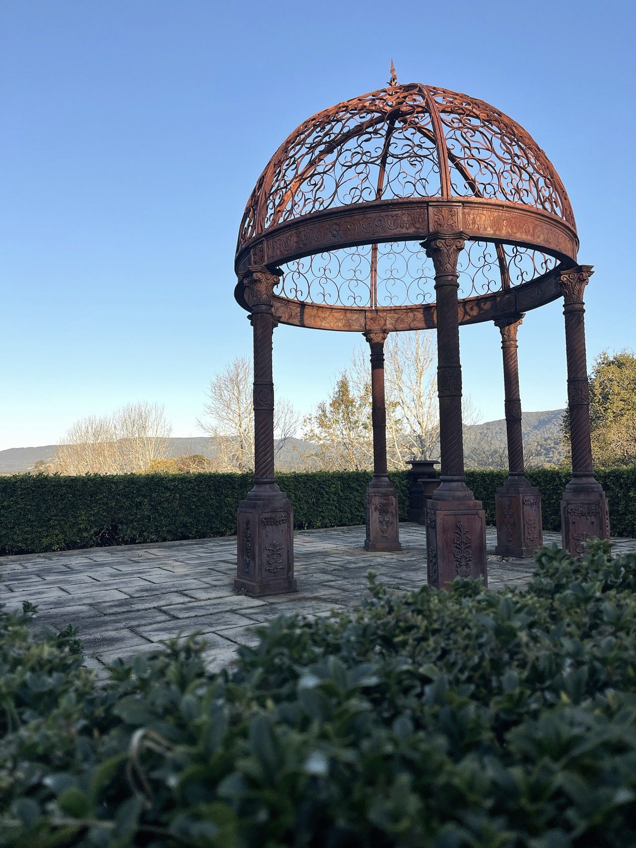 En pavillon med kuppelformet tag og detaljeret metaludsmykning, omgivet af en brostensbelagt plads og en hæk.